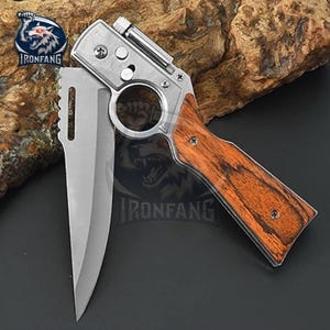 May include: A folding knife with a stainless steel blade and a handle designed to resemble a pistol. The handle features a wood grain pattern. The knife is open, displaying the blade. The brand logo "IRONFANG" is visible.