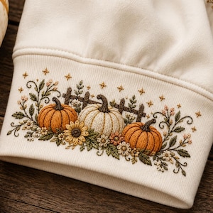 May include: A cream-colored sweatshirt cuff featuring detailed embroidery of two orange pumpkins and one white pumpkin, a wooden fence, sunflowers, and small green leaves. The design is accented with tiny gold star shapes scattered above the pumpkins.