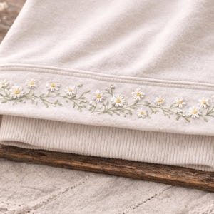 Può includere: Felpa color crema con dettaglio a catena di margherite ricamate lungo lo scollo. Il ricamo presenta margherite bianche con centri gialli e steli verdi. La felpa è piegata su una superficie di legno.