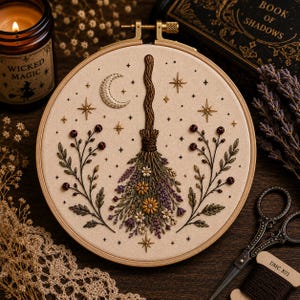 May include: A circular wooden embroidery hoop containing a fabric panel with a hand-stitched design of a broomstick adorned with flowers and lavender. The scene includes a crescent moon, stars, and two symmetrical floral branches on the sides.