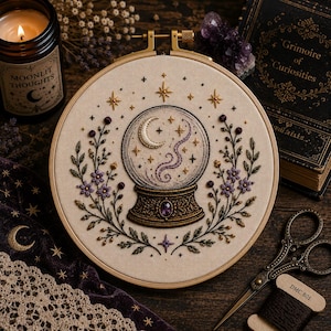 May include: A circular embroidery hoop featuring a stitched crystal ball design on cream fabric. The crystal ball contains a crescent moon and swirling purple smoke, flanked by floral vines with purple flowers and gold star accents.