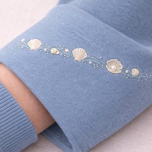 May include: Close-up of a light blue knit sleeve with embroidered seashells, pearls, and a small star. The cuff is ribbed, and the embroidery is in cream and white. The sleeve is folded over, revealing the arm.