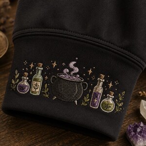 May include: A black sweatshirt sleeve cuff featuring detailed embroidery of a central black cauldron with purple steam, flanked by four glass potion bottles in shades of green and purple, accented with small embroidered stars and green foliage.