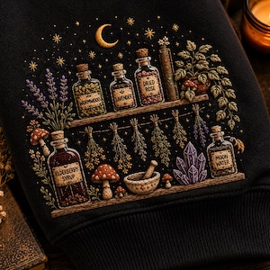 Puede incluir: Una sudadera negra con un gráfico detallado de estantes con botellas de vidrio etiquetadas como Wormwood, Lavender, Dried Rose, Elderberry Syrup y Moon Water. El diseño incluye hierbas colgantes, setas, un mortero y cristales.