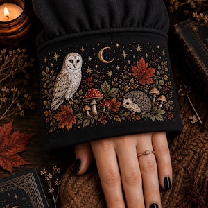 May include: A black long-sleeved garment featuring an embroidered cuff with a detailed nature scene. The design includes a white owl, a hedgehog, red mushrooms, and autumn leaves under a crescent moon and stars, all rendered in warm earth tones.