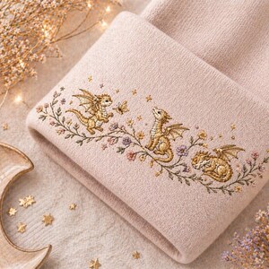 May include: A pale pink beanie featuring an embroidered design of three golden dragons, butterflies, and flowers. The dragons are in different poses, with one sleeping. The embroidery is detailed and features gold, green, and purple colours.