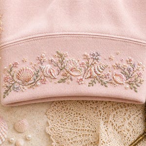 May include: Pale pink sweatshirt with embroidered seashells and floral details. The embroidery includes seashells, flowers, and pearls in pink, green, and cream. The design is on the bottom hem of the sweatshirt.