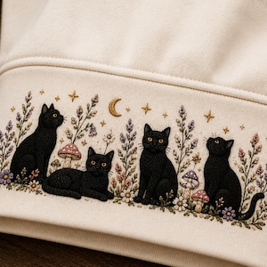 May include: A cream-coloured sweatshirt sleeve cuff featuring detailed embroidery of four black cats sitting among colourful wildflowers, mushrooms, and a golden crescent moon with small stars scattered across the fabric above the cats.