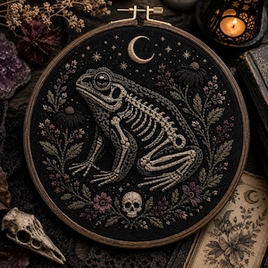 May include: A circular embroidery hoop containing black fabric with a detailed thread-stitched design of a toad skeleton. The toad is surrounded by embroidered flowers, leaves, a crescent moon, stars, and a small skull at the bottom centre.