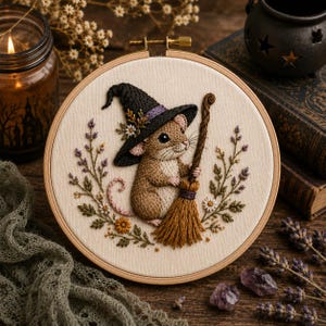 May include: A circular embroidery hoop featuring a tan mouse wearing a black pointed hat and holding a brown broom. The mouse is surrounded by a wreath of embroidered lavender sprigs, small white flowers, and green leaves on cream fabric.