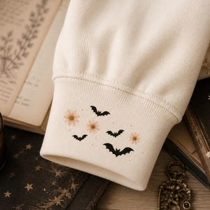 May include: A cream-colored long-sleeve sweatshirt cuff featuring a small embroidered design of three black bats and three light pink daisy flowers, with tiny scattered dots surrounding the motifs on the ribbed fabric sleeve end.