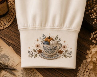 Puede incluir: Una manga de sudadera color crema con un bordado detallado de una taza de té floral llena de tres pequeñas calabazas en naranja, azul y blanco. La taza está rodeada de hojas delicadas, flores pequeñas y pequeños detalles de estrellas.