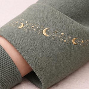 Puede incluir: Primer plano de una manga verde salvia con un diseño celestial bordado en oro. El diseño presenta tres lunas crecientes y varias estrellas. El puño es acanalado y la manga está hecha de una tela suave y texturizada.