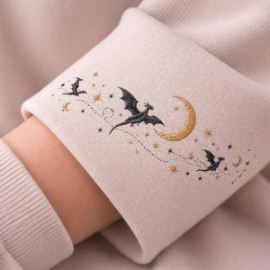 Peut inclure: Gros plan sur une manche de sweat-shirt beige avec des broderies de dragons, d'étoiles et d'un croissant de lune. Le motif présente des dragons noirs et des étoiles et une lune dorées, créant un thème céleste.