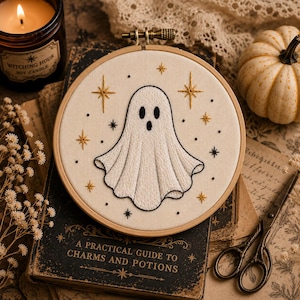 Puede incluir: Un bastidor de bordado circular de madera que contiene un panel de tela blanca con un diseño de fantasma bordado a mano. El fantasma es blanco con contornos y rasgos faciales negros, rodeado de estrellas doradas y negras sobre fondo crema.