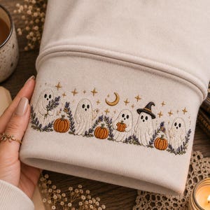 Puede incluir: Una manga de sudadera beige con un bordado detallado de cinco fantasmas blancos, calabazas naranjas, ramas de lavanda morada y una luna creciente dorada con pequeñas estrellas. Los fantasmas tienen ojos y bocas de color negro.