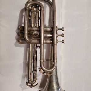 May include: A vintage silver-colored cornet with a flared bell. The instrument has three valves, a mouthpiece, and intricate tubing. The cornet shows signs of age and wear, with a tarnished finish. The bell has a decorative design.