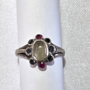 May include: A silver ring featuring an oval, pale green gemstone at the center, surrounded by smaller gemstones in dark blue, black, and red. The ring's band has a textured design. Jewelry.