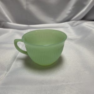 May include: A pale mint green glass teacup featuring a translucent finish and a delicate vertical ribbed pattern along the exterior. The cup has a rounded bowl shape and a small, curved handle extending from the side, crafted from smooth glass.