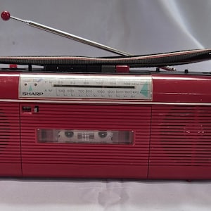 May include: A rectangular red Sharp portable radio and cassette player featuring a horizontal tuning dial, a silver telescopic antenna with a red tip, a black carrying strap, and a central cassette compartment with horizontal speaker grilles.