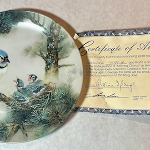 May include: A decorative plate featuring a blue jay with three chicks in a nest, set against a backdrop of pine branches. The plate is accompanied by a certificate of authenticity. The plate is part of the "Morning Chorus" collection.