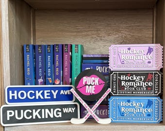 May include: A shelf display featuring books by Rachel Reid, hockey-themed decor, and book club tickets. Items include a hockey puck with "Puck Me" text, hockey stick, and street signs reading "Hockey Ave" and "Pucking Way". Tickets read "Hockey Romance Book Club".