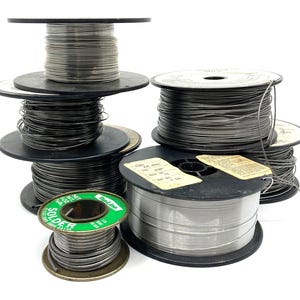 May include: A collection of solder wire spools in various sizes. The spools are black with silver wire wound around them. One spool has a green label with the word "SOLDER" in white. Some spools have attached labels.