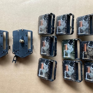 May include: A collection of eleven quartz clock movement mechanisms. Two units feature black plastic housings with brass-colored shafts, while nine units have transparent plastic casings revealing internal gears, copper coils, and white components.