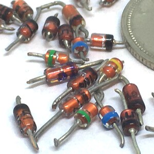 May include: A collection of small, brown, glass diodes with colored bands. The diodes have silver leads and are scattered on a white surface.