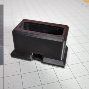 May include: A black, rectangular object with a hollow interior, possibly a magazine holder or similar item. The object has a square base and a slightly textured surface, sitting on a grid-patterned surface.