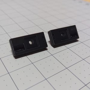May include: Two small, rectangular black plastic components with a hole and a slot. One component has the letters "SAT" printed on it. The components are on a light gray surface with a grid pattern.