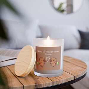 May include: A lit, frosted glass candle with a wooden lid sits on a wooden table. The candle has a label with the text "Thanks for being my fartner forever" and a cartoon illustration of two brown animals. The candle is a home decor item.