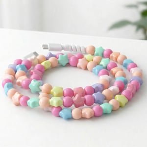May include: A decorative charging cable featuring pastel-coloured beads. The cable is decorated with round and star-shaped beads in pink, blue, yellow, and peach. The cable is white and has a USB-C connector.