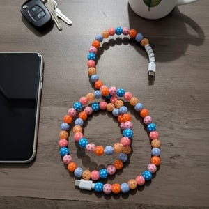 May include: A decorative USB-C charging cable featuring multi-coloured beads in pink, orange, blue, and peach. A white connector is attached. A smartphone, keys, and a mug with a leaf design are also visible on a wooden surface.