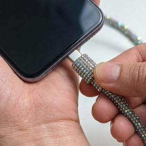 以下が含まれることがあります： 装飾的なUSB-Cケーブルで充電されているスマートフォン。ケーブルは虹色のラインストーンで覆われており、電話は黒です。人の手はケーブルと電話を持っています。