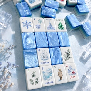 Puede incluir: Una colección de fichas de mahjong rectangulares que presentan una mezcla de patrones de mármol azul sólido y fichas blancas con ilustraciones impresas de copos de nieve, flores, cristales y textos como SOAP, JOKER, GREEN, FLOWER y SOUTH.