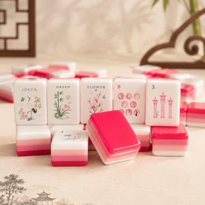 Op de afbeelding: Een set witte mahjongtegels met roze gradi&euml;ntvoeten. De tegels tonen diverse illustraties waaronder een kraanvogel, bamboe, bloemen en pagodes, met tekstlabels zoals JOKER, GREEN, FLOWER 5, 8 en 3 gedrukt in zwarte of roze inkt.