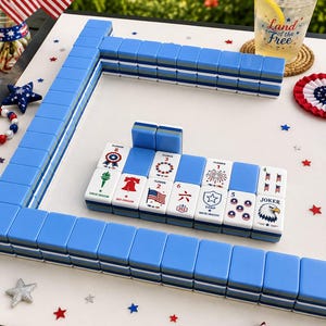 May include: A set of blue and white mahjong tiles arranged on a surface with patriotic decorations. The tiles feature various designs including a bald eagle, the Liberty Bell, fireworks, and American flags, with some tiles labeled as Flower or Joker.