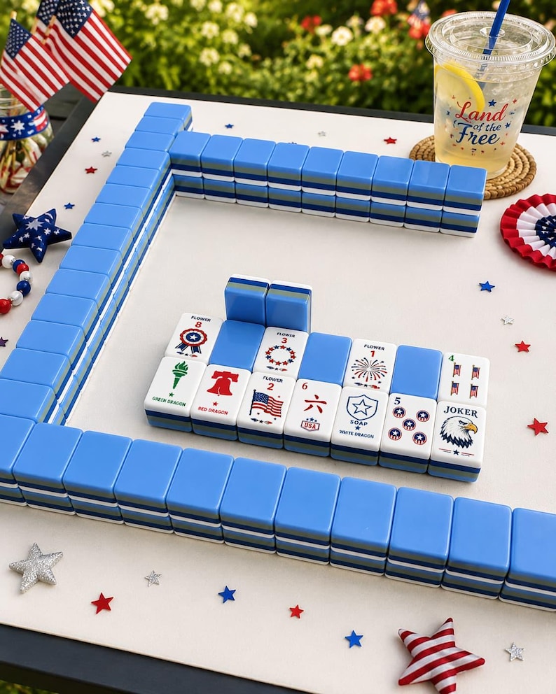 May include: A set of blue and white mahjong tiles arranged on a surface with patriotic decorations. The tiles feature various designs including a bald eagle, the Liberty Bell, fireworks, and American flags, with some tiles labeled as Flower or Joker.