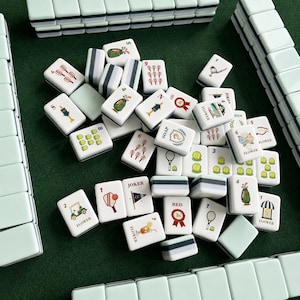 May include: A set of rectangular mahjong tiles with a pale green and white color scheme. The tiles feature various colorful illustrations including sports equipment, flowers, and text such as JOKER, SOAP, RED, and FLOWER, arranged on a green surface.