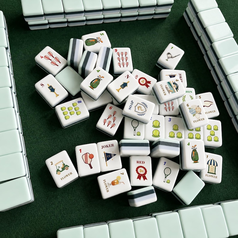 May include: A set of rectangular mahjong tiles with a pale green and white color scheme. The tiles feature various colorful illustrations including sports equipment, flowers, and text such as JOKER, SOAP, RED, and FLOWER, arranged on a green surface.