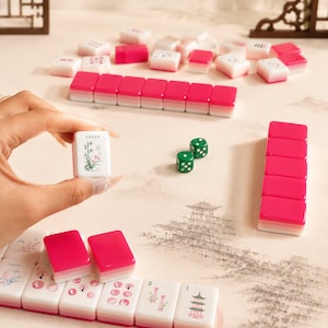 May include: A Mahjong set featuring white tiles with pink and green decorative illustrations and text, including a tile labeled GREEN with a bamboo design. The set includes two green dice and several rows of tiles with pink backs and white fronts.