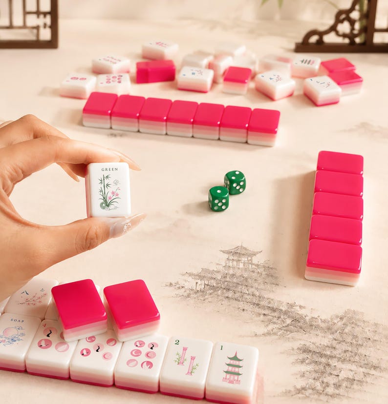 May include: A Mahjong set featuring white tiles with pink and green decorative illustrations and text, including a tile labeled GREEN with a bamboo design. The set includes two green dice and several rows of tiles with pink backs and white fronts.