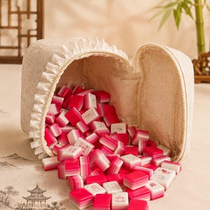 May include: A beige fabric zippered pouch with a ruffled trim edge, filled with a collection of small rectangular mahjong tiles. The tiles feature a gradient design transitioning from white to pink and deep red, with some displaying intricate illustrations.