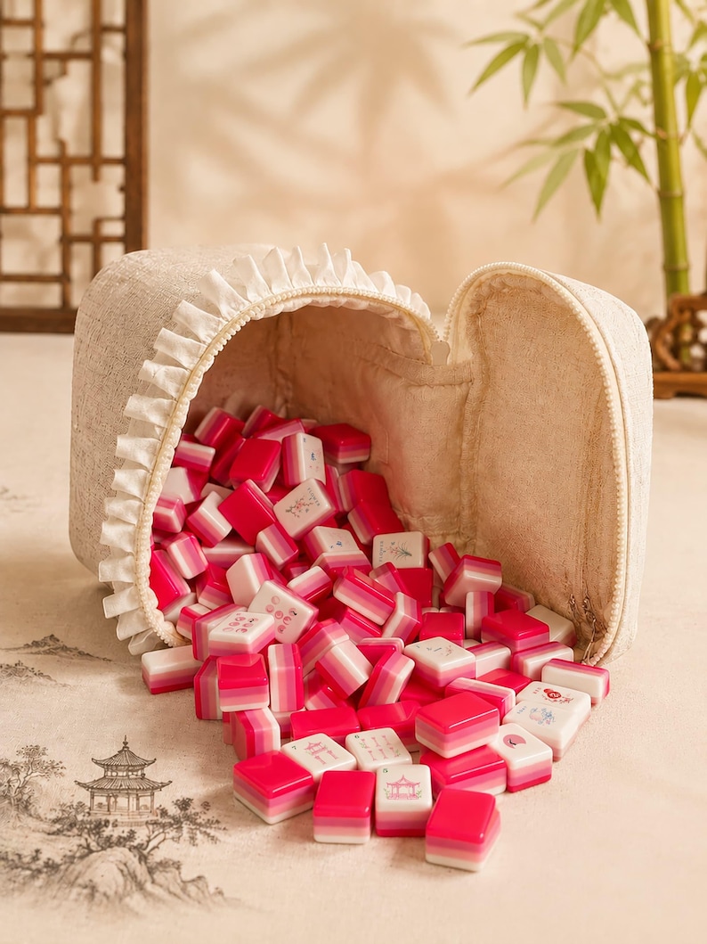 May include: A beige fabric zippered pouch with a ruffled trim edge, filled with a collection of small rectangular mahjong tiles. The tiles feature a gradient design transitioning from white to pink and deep red, with some displaying intricate illustrations.