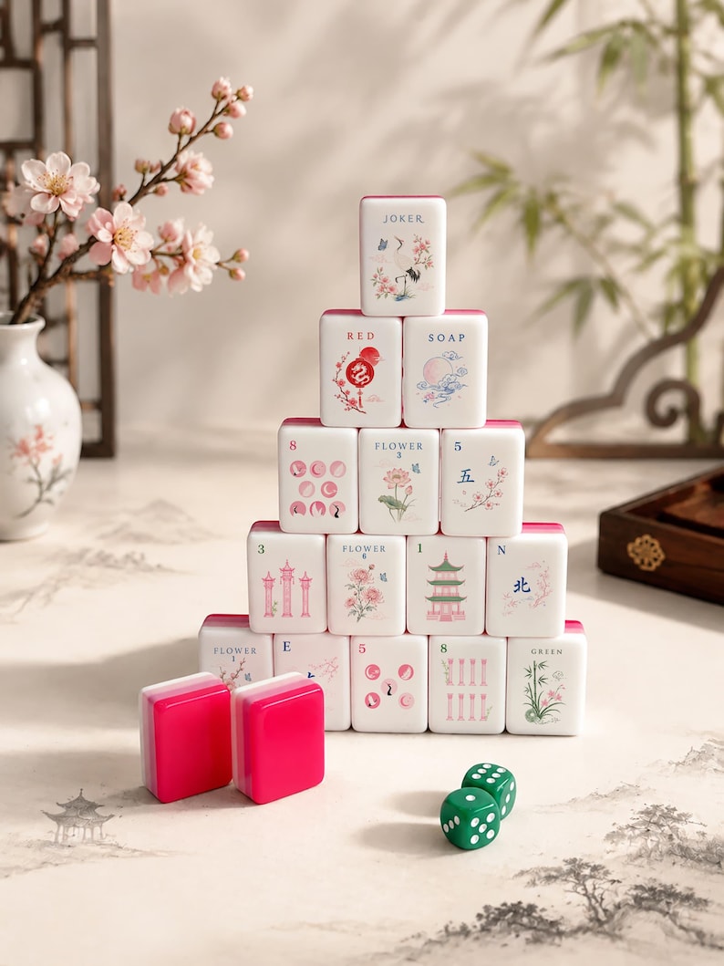 May include: A pyramid of white mahjong tiles with pink backs, featuring various illustrations including flowers, pagodas, and text like JOKER, RED, SOAP, and FLOWER. Two solid pink tiles and two green dice with white dots are placed in the foreground.