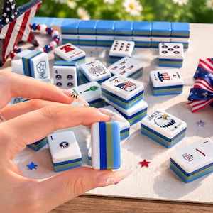 May include: A set of white mahjong tiles featuring patriotic American-themed graphics including eagles, flags, and stars. The tiles have blue and green striped sides and are arranged with a star-shaped decoration and red, white, and blue ribbons.