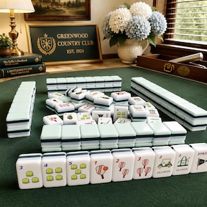 May include: A set of mahjong tiles featuring golf-themed illustrations, including golf balls, clubs, carts, and flags. The tiles are white with light green and dark green accents, arranged in rows and stacks on a dark green felt surface.