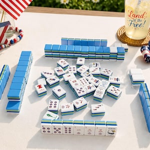 May include: A Mahjong set featuring white tiles with blue, green, and yellow layered bases. The tiles display various patriotic American graphics, including flags, eagles, and fireworks. Some tiles are arranged in rows, while others are scattered.