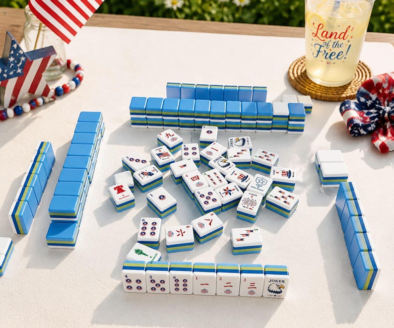 May include: A Mahjong set featuring white tiles with blue, green, and yellow layered bases. The tiles display various patriotic American graphics, including flags, eagles, and fireworks. Some tiles are arranged in rows, while others are scattered.